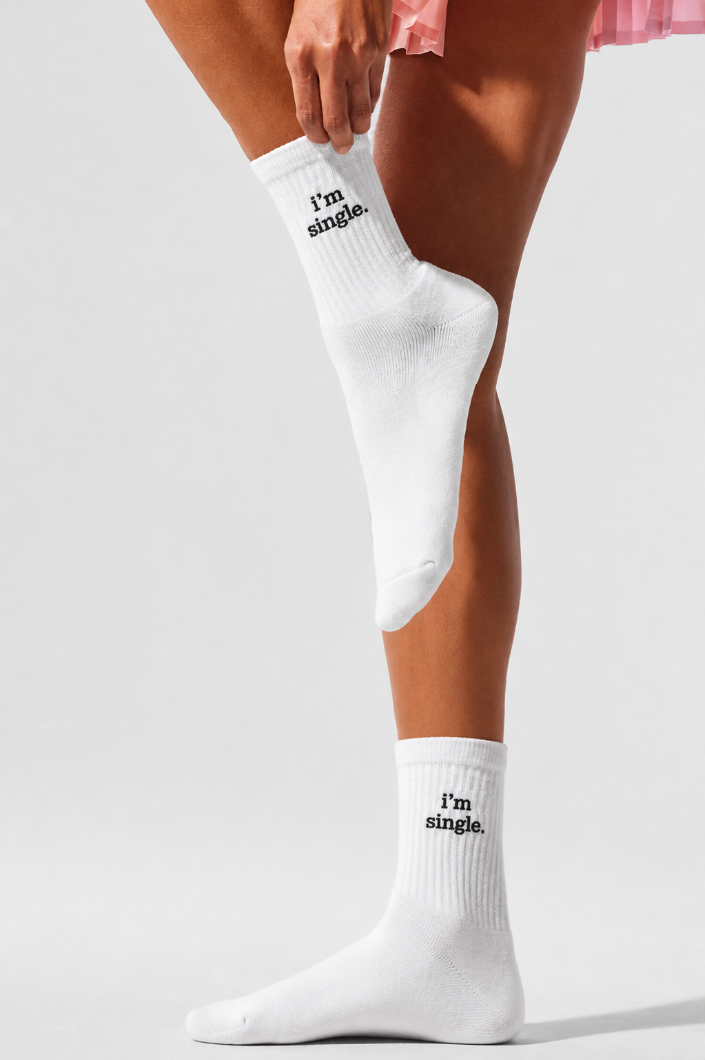 A person in a pink skirt pulling up a white ribbed crew sock embroidered with 'i'm single.' in black, photographed against a white studio background.