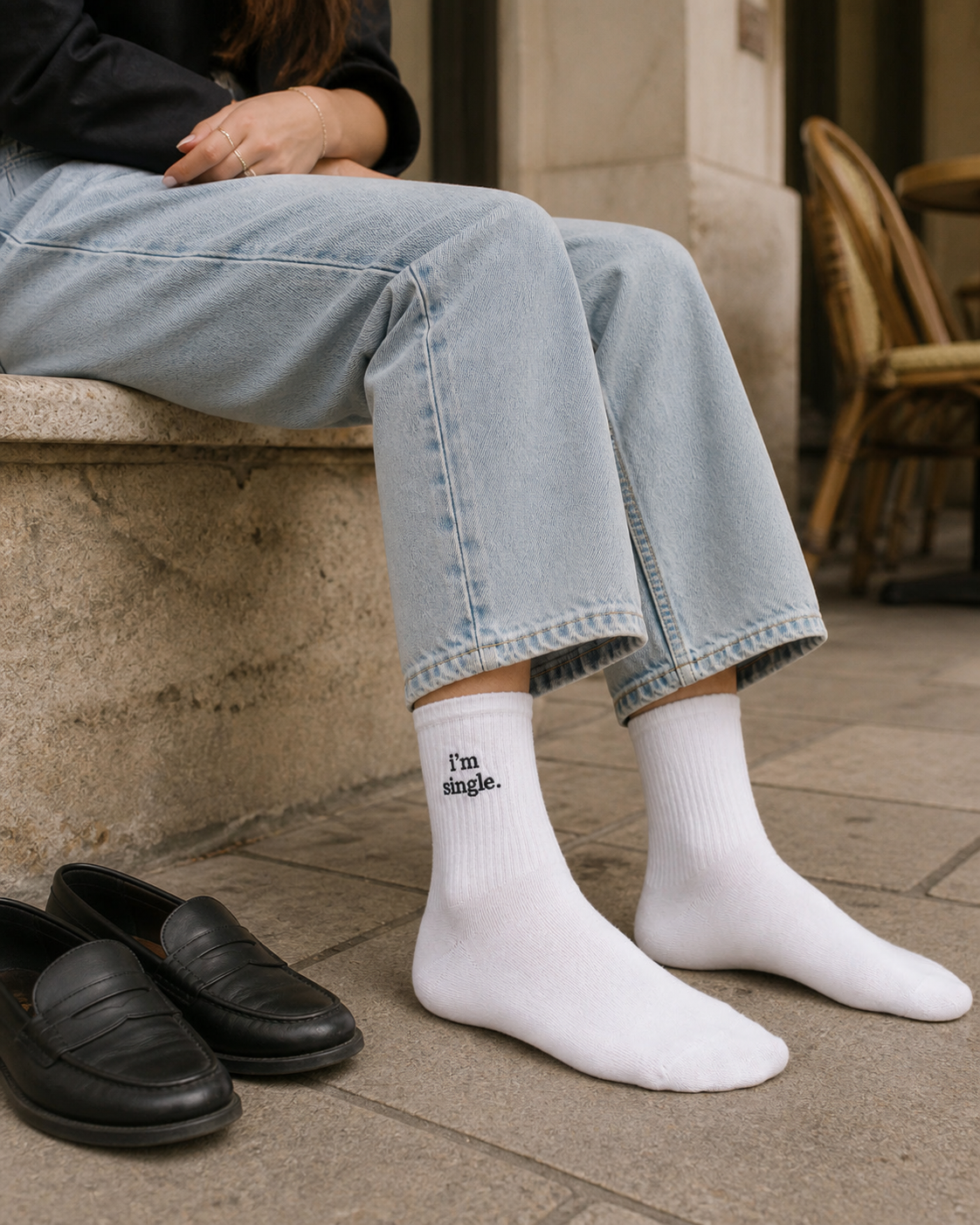 A person sitting on a stone ledge outside a cafe in light-wash jeans and white ribbed crew socks embroidered with 'i'm single.', with black loafers on the pavement beside them.