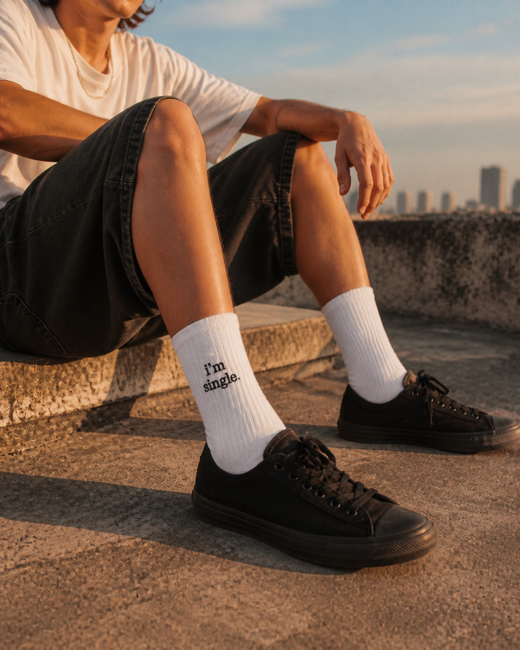 A man sitting on a rooftop at golden hour in black shorts and white ribbed crew socks embroidered with 'i'm single.', worn with black low-top sneakers.