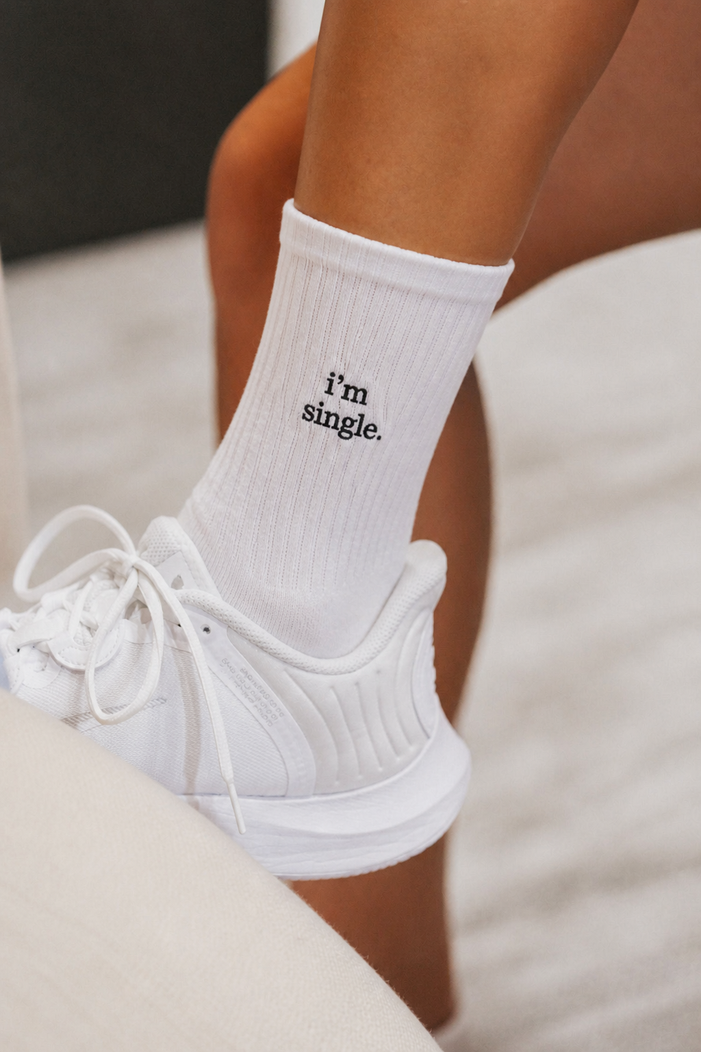 Close-up of a white ribbed crew sock embroidered with 'i'm single.' in black, worn with a white sneaker.