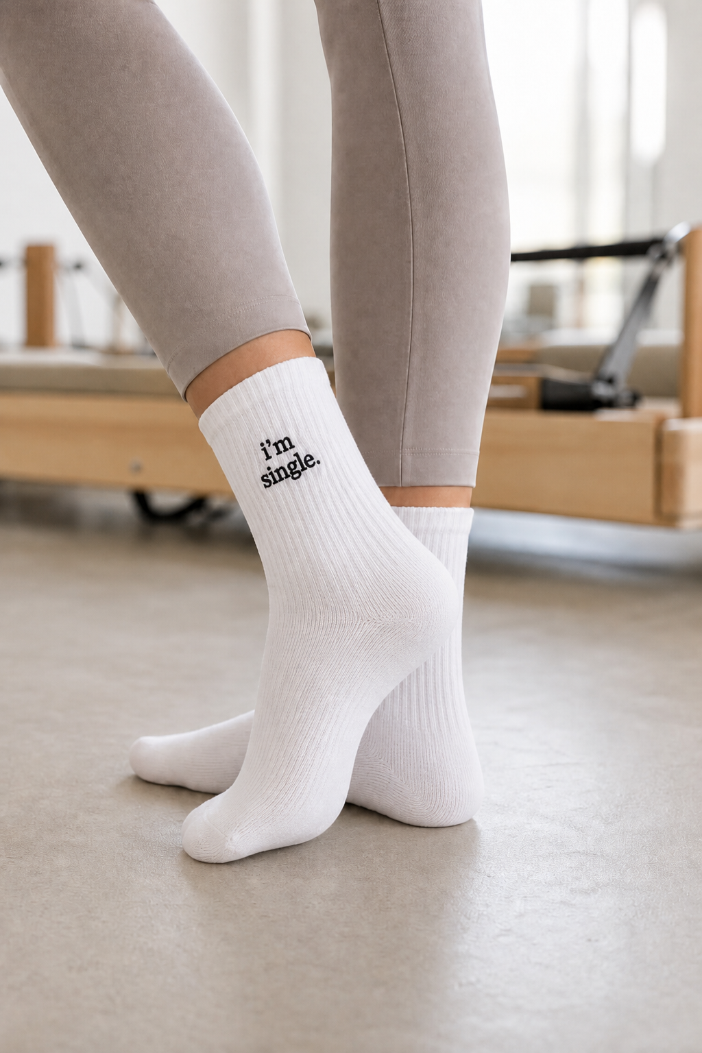 A person in grey leggings wearing white ribbed crew socks embroidered with 'i'm single.' in black, standing in a pilates studio.