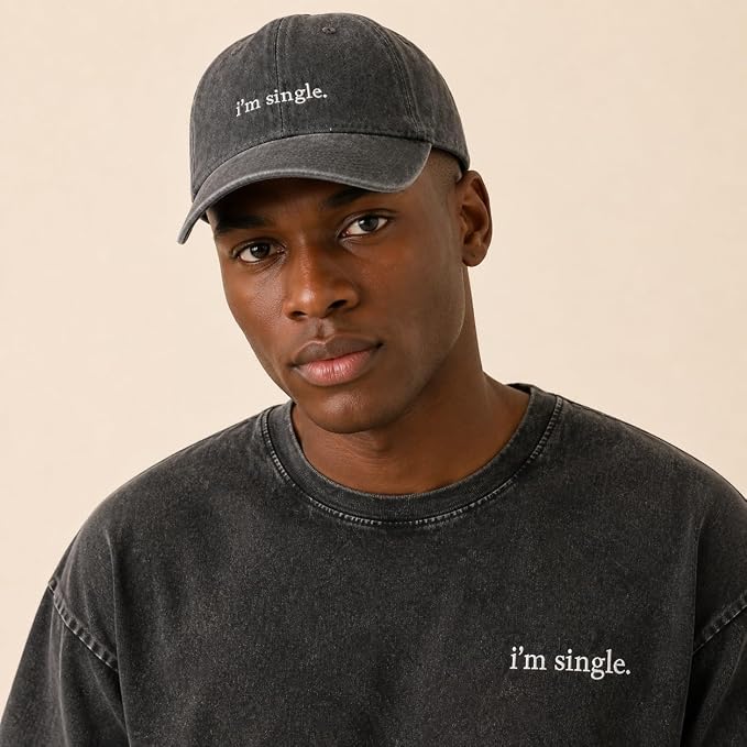 A man wearing a dark gray washed baseball cap and matching dark gray washed t-shirt, both featuring the white embroidered text 'i'm single.' on the front, against a solid light beige background.