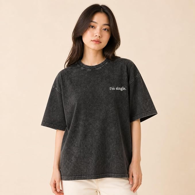 A woman wearing an oversized dark gray acid-wash t-shirt with the text 'I'm single.' printed in white on the left chest, paired with cream-colored pants, set against a solid beige background.