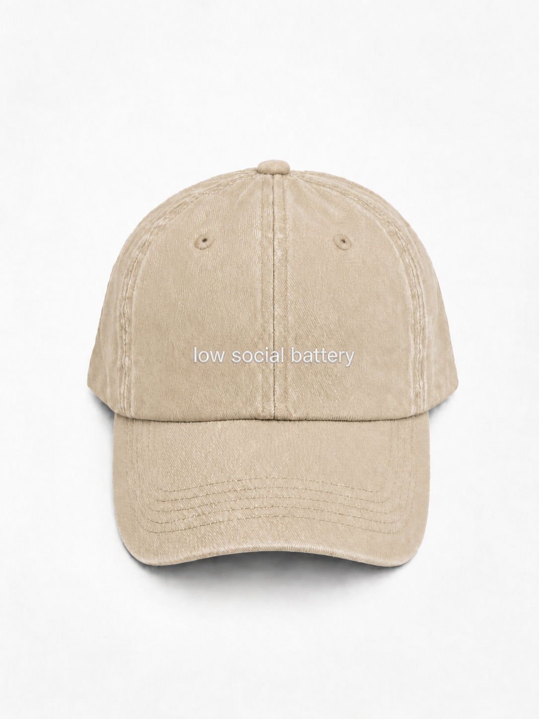 A beige washed cotton baseball cap with the phrase 'low social battery' embroidered in white across the front, set against a plain white background.