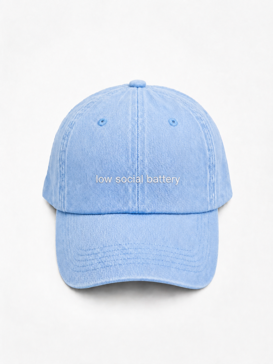 A light blue washed cotton baseball cap with the phrase 'low social battery' embroidered in white across the front, set against a plain white background.