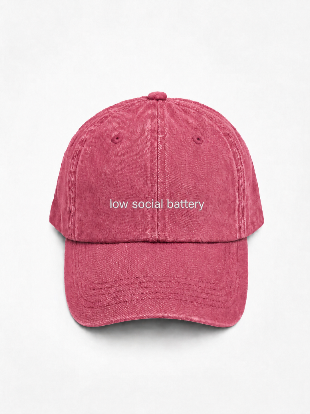 A washed pink baseball cap with the phrase 'low social battery' embroidered in white across the front, shown against a plain white background.