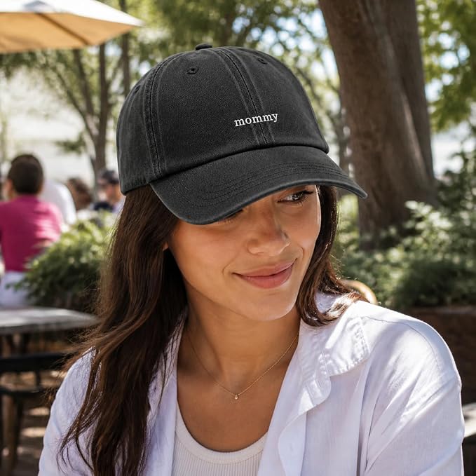 A woman wearing a black baseball cap with the word 'mommy' embroidered in white on the front, photographed outdoors at a cafe or park setting.