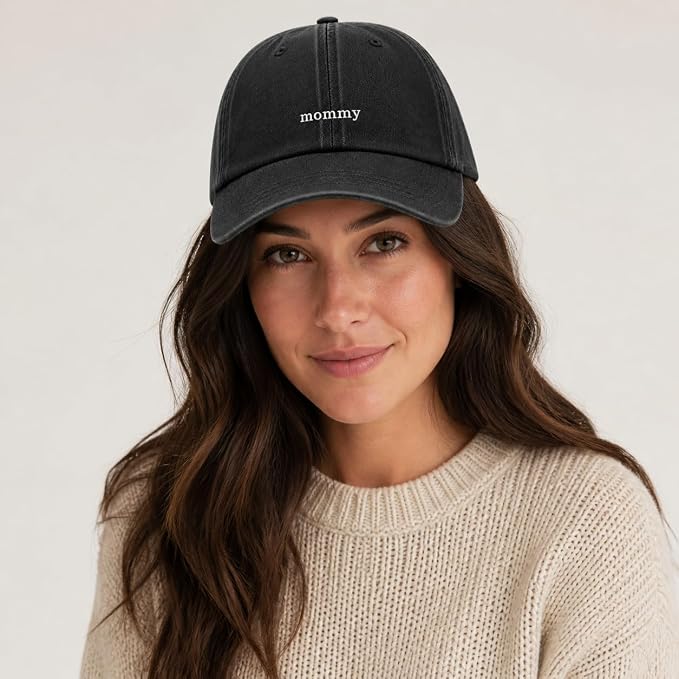 A woman wearing a black washed baseball cap with the word 'mommy' embroidered in white on the front, paired with a cream knit sweater against a light gray background.