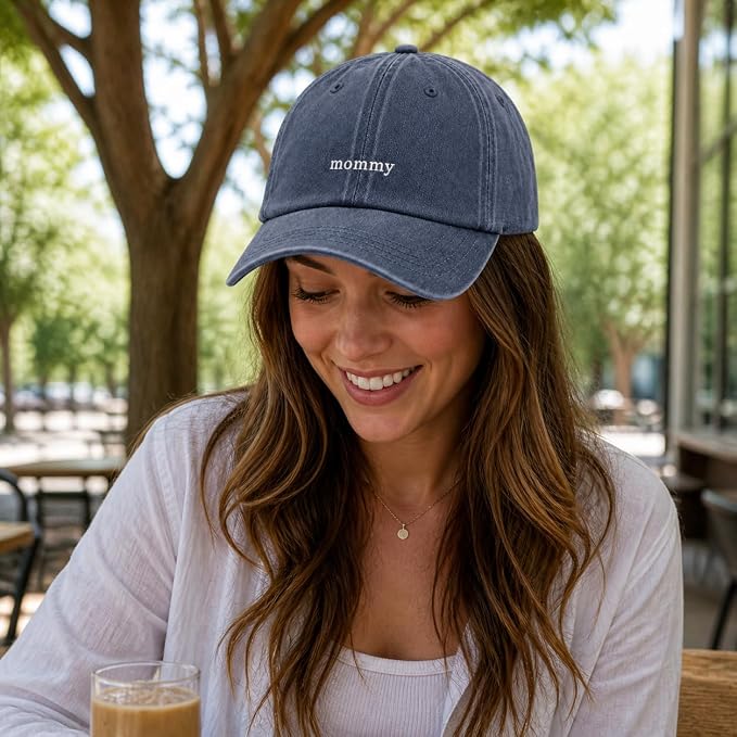 A woman wearing a navy blue baseball cap with 'mommy' embroidered in white on the front, sitting at an outdoor cafe table.