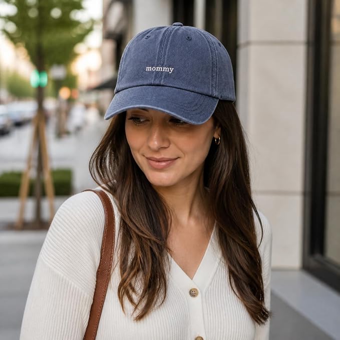 A woman wearing a blue denim baseball cap with 'mommy' embroidered in white on the front, photographed outdoors on a city street.