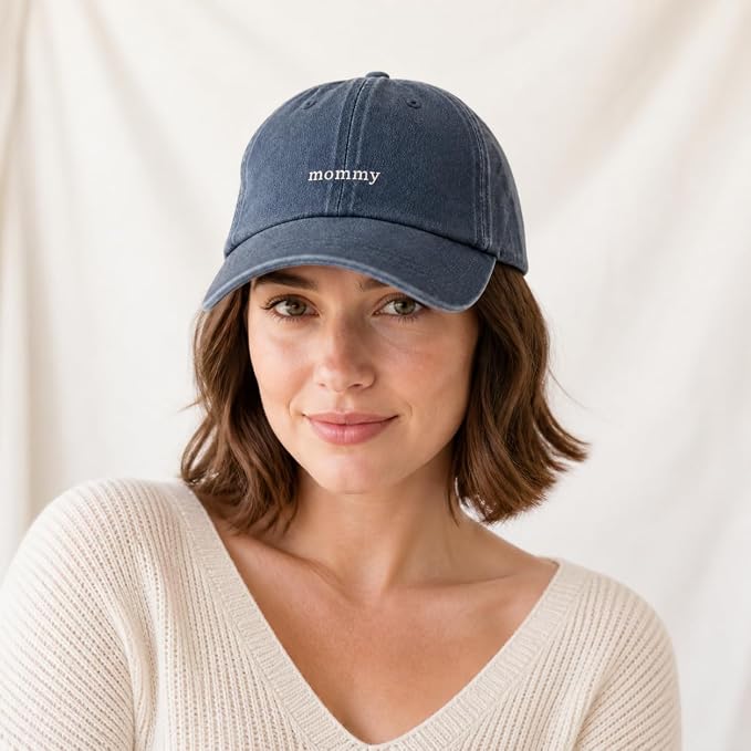 A woman wearing a navy blue distressed baseball cap with the word 'mommy' embroidered in white on the front, photographed against a soft cream fabric backdrop.
