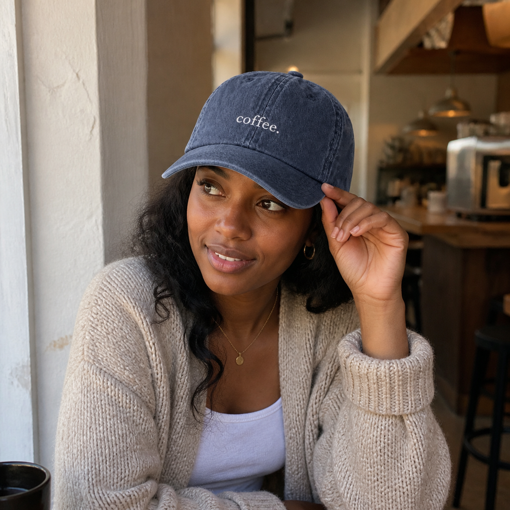 A woman in an oatmeal cardigan adjusting the brim of the washed navy 'coffee.' cap inside a cafe.