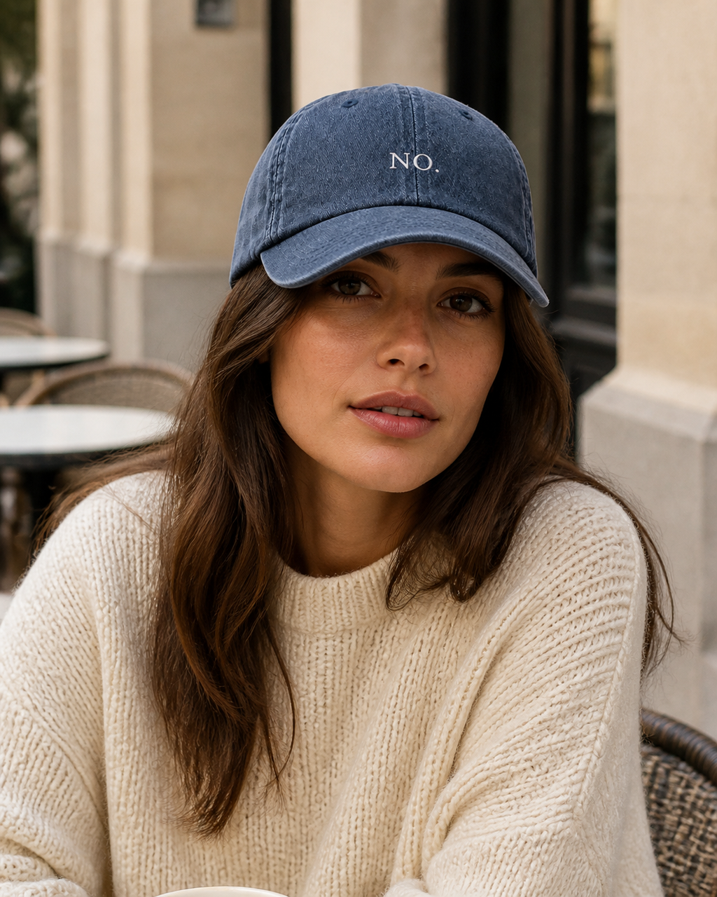 A woman in a cream knit sweater wearing the washed navy 'NO.' cap at an outdoor cafe table.