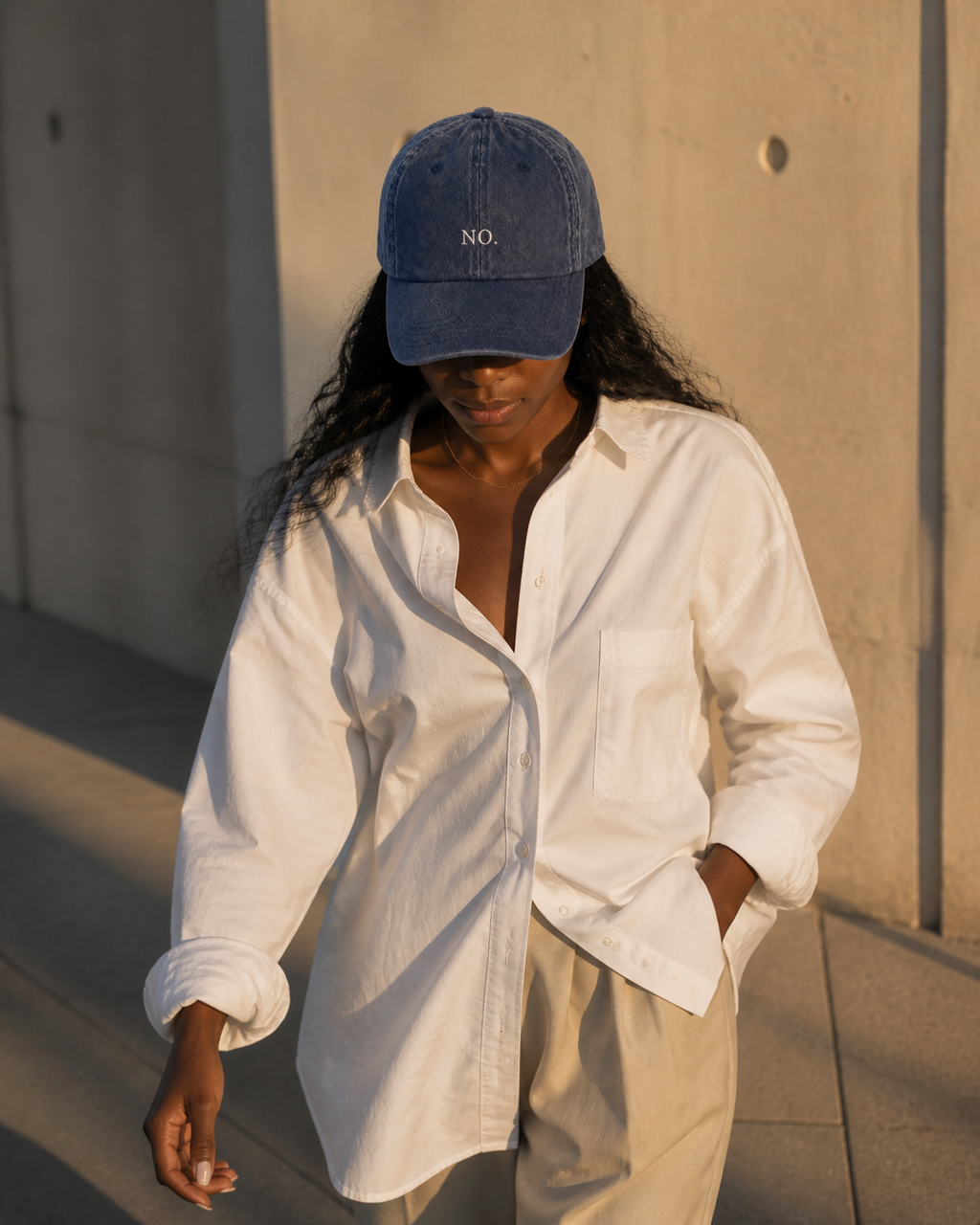 A woman in a white linen shirt wearing the washed navy 'NO.' cap, walking past a concrete wall in golden evening light.