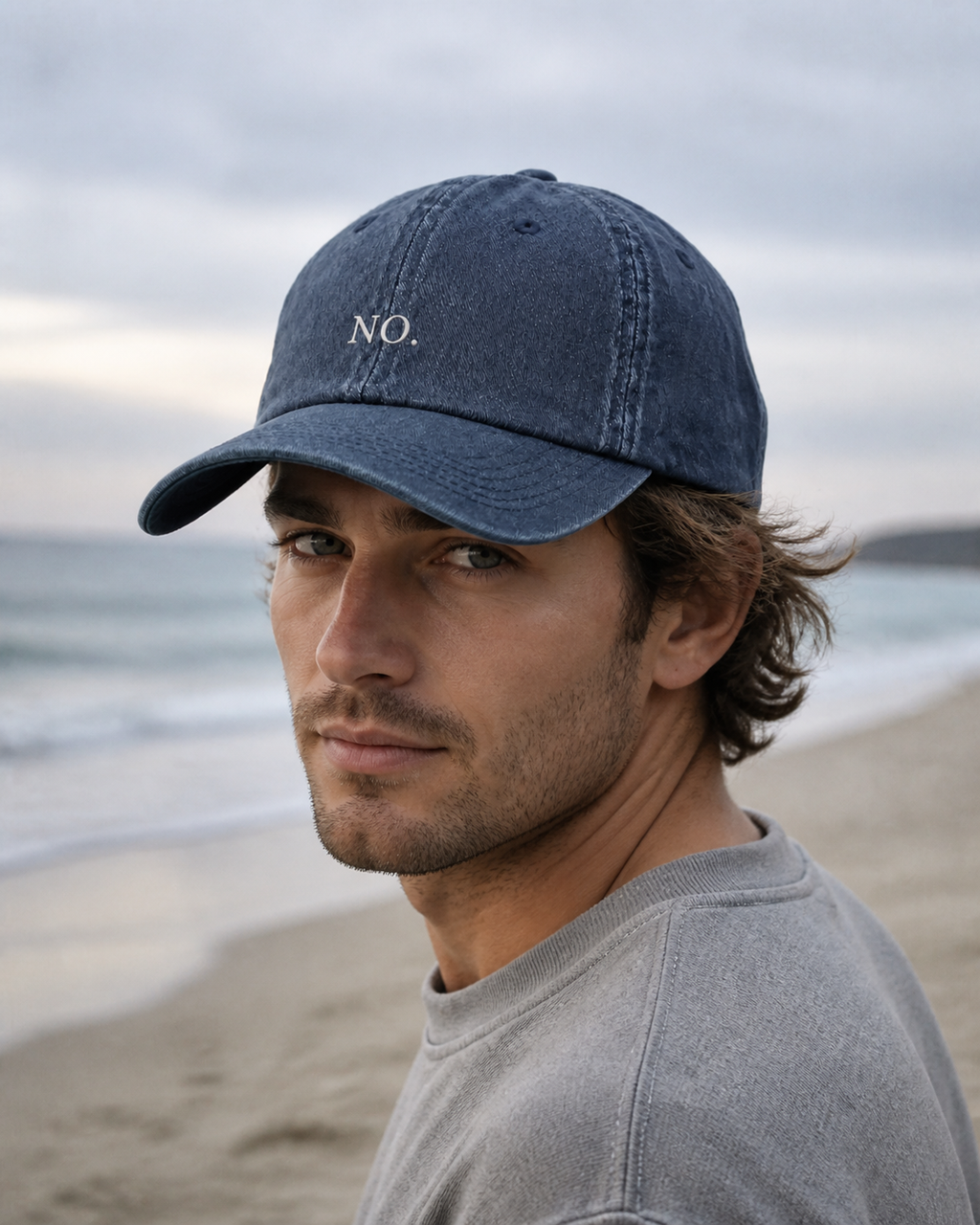 A man in a grey sweatshirt wearing the washed navy 'NO.' cap on a beach at dusk.