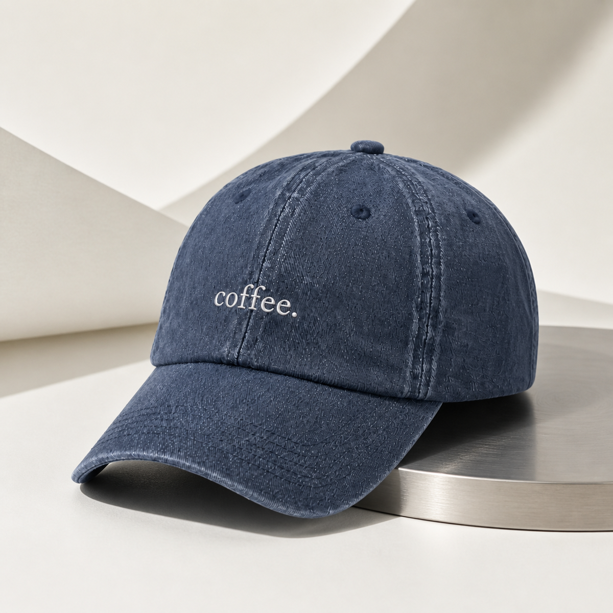 The washed navy 'coffee.' cap on a steel disc against a sculptural cream backdrop.