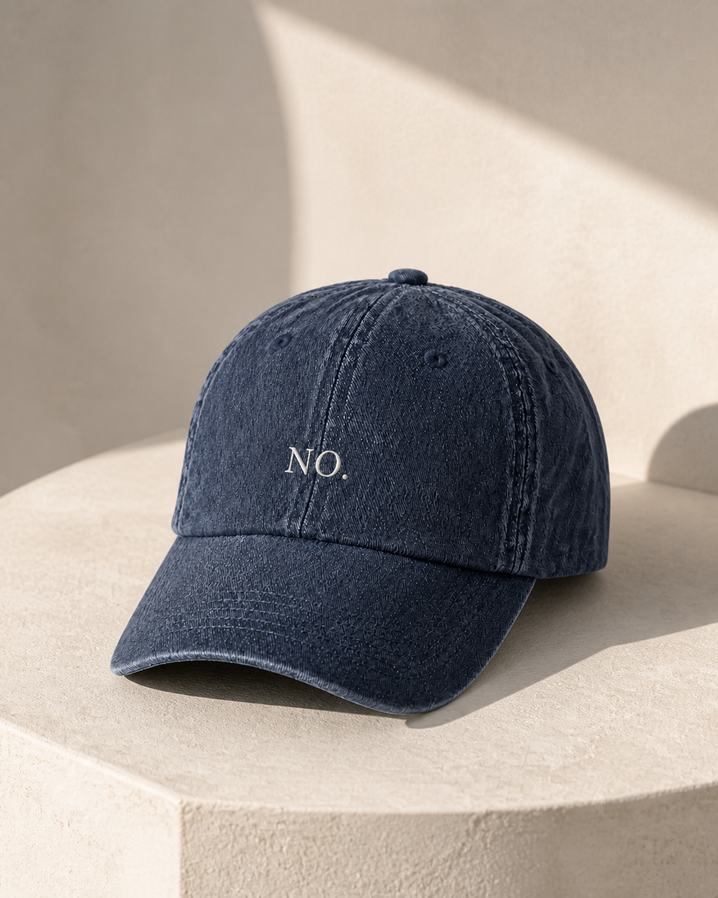 The washed navy 'NO.' cap on a pale stone plinth in warm directional sunlight.
