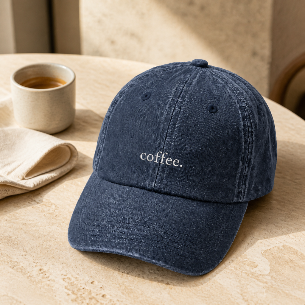 The washed navy 'coffee.' cap on a round travertine table beside an espresso and a linen napkin.