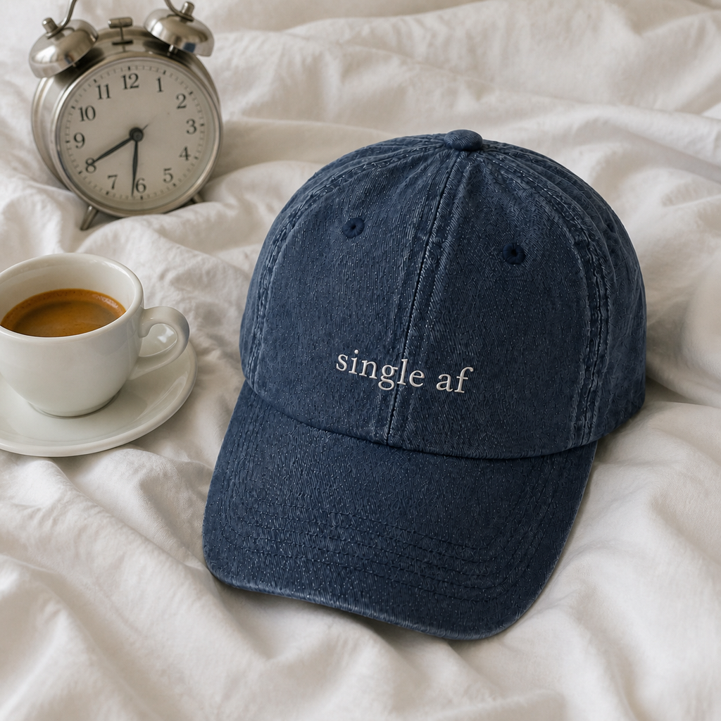 A navy baseball cap embroidered with 'single af' in white, resting on rumpled bedding in morning light.