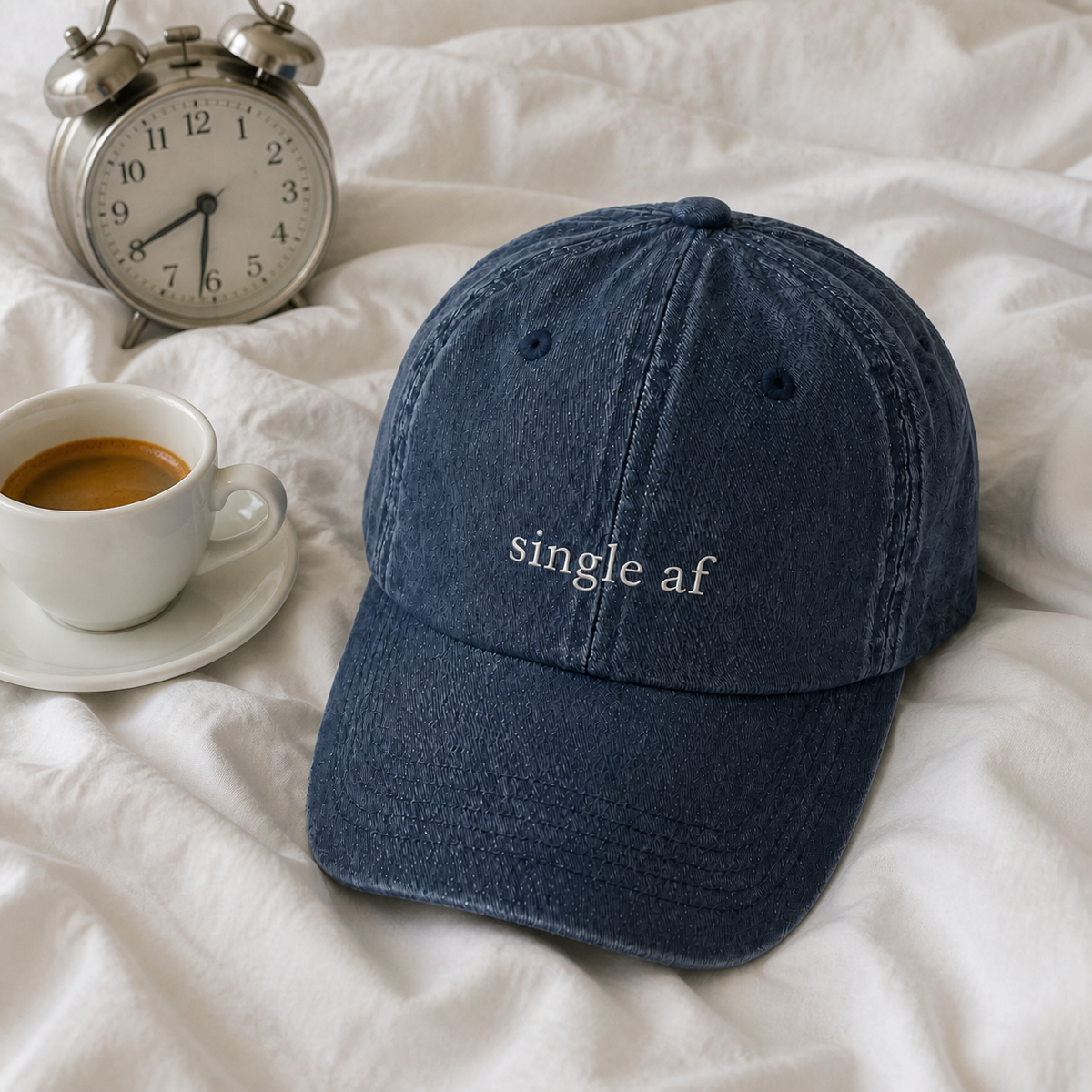 A navy baseball cap embroidered with 'single af' in white, resting on rumpled bedding in morning light.