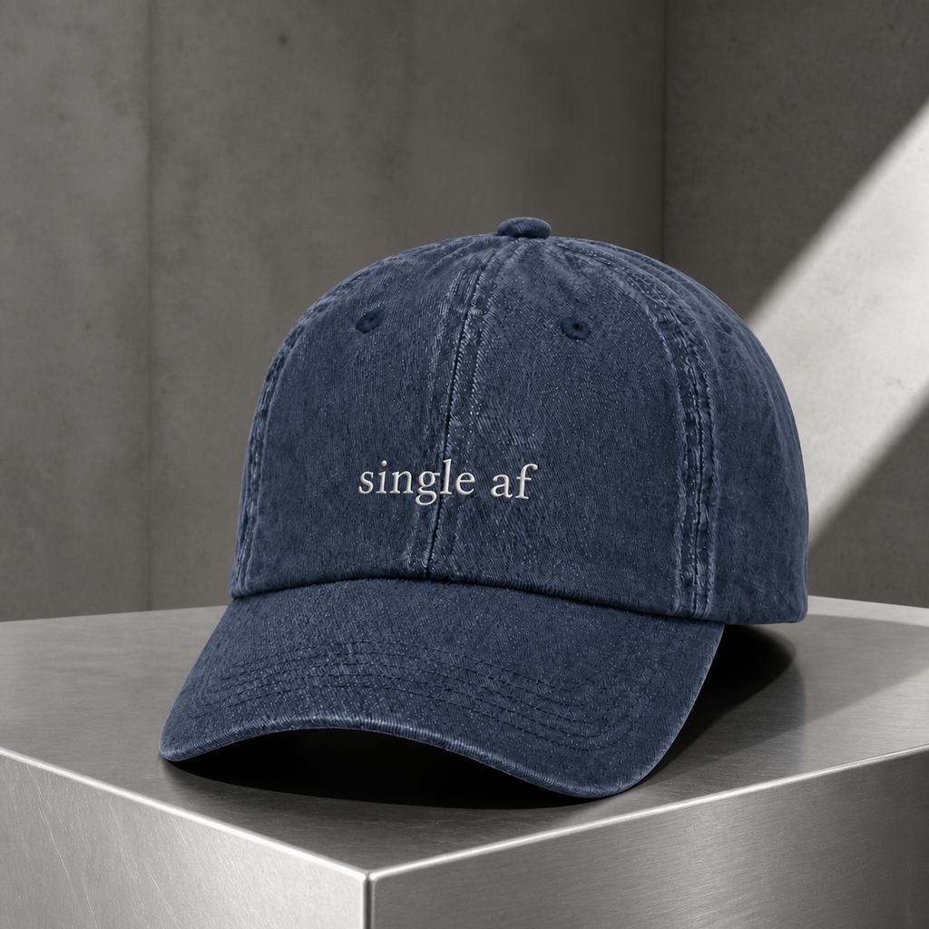 A navy baseball cap embroidered with 'single af' in white, photographed on a stainless steel surface in a studio.