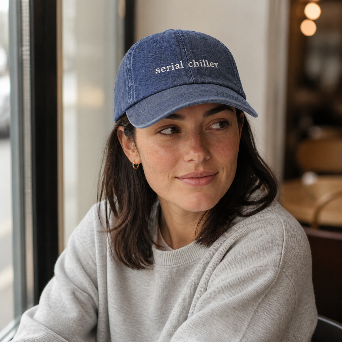 A person wearing a navy baseball cap embroidered with 'serial chiller' in white, seated in a cafe.