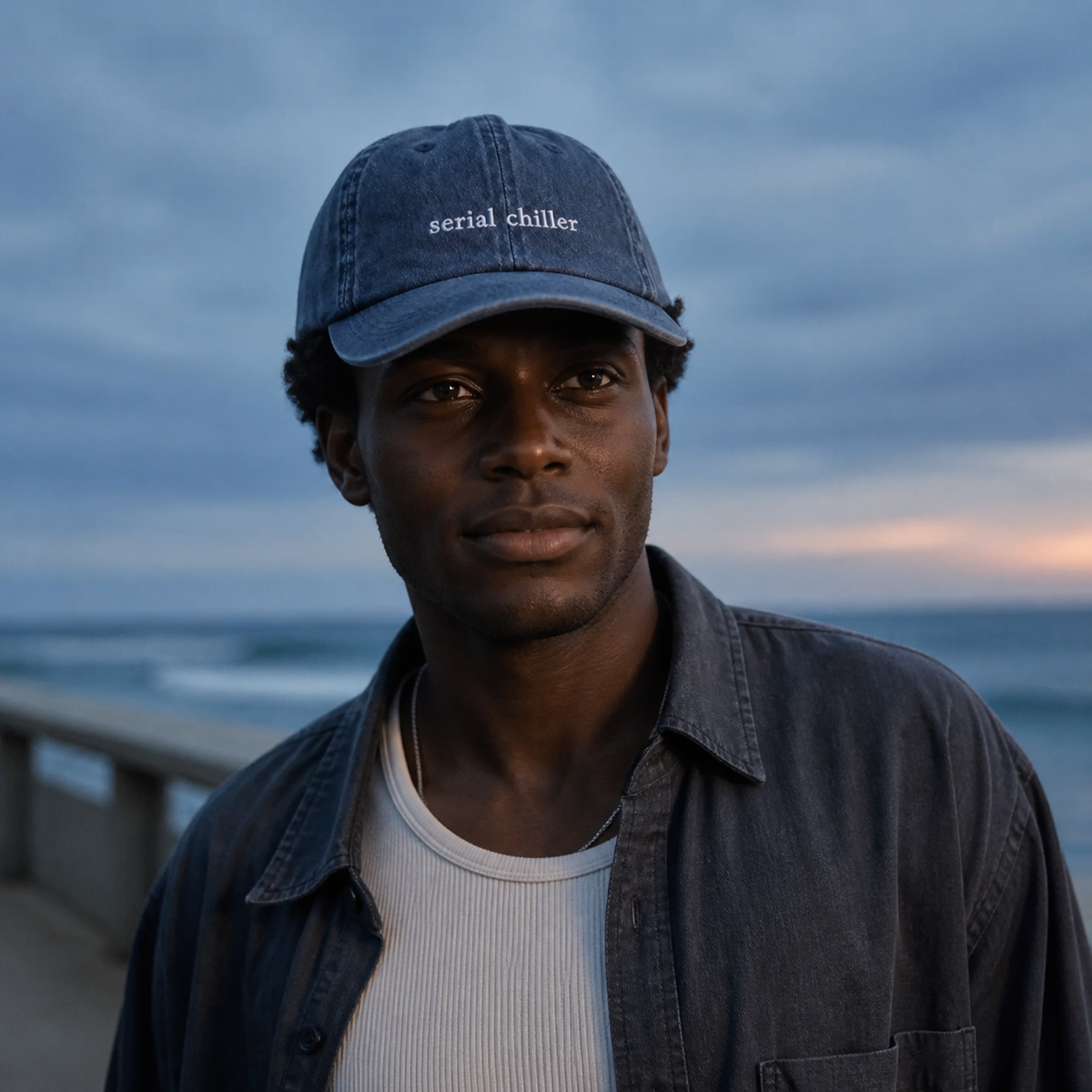 A person wearing a navy baseball cap embroidered with 'serial chiller' in white by the sea.