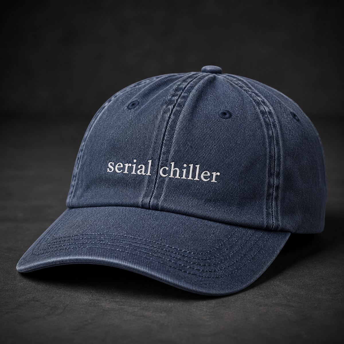 A navy baseball cap embroidered with 'serial chiller' in white, photographed against a dark studio background.