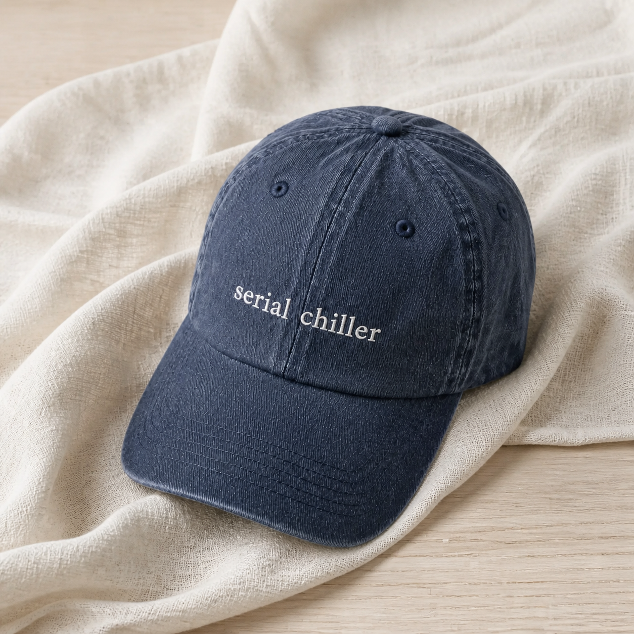 A navy baseball cap embroidered with 'serial chiller' in white, resting on draped linen.