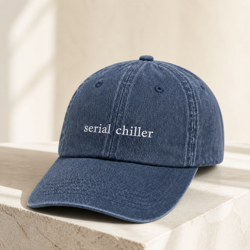 A navy baseball cap embroidered with 'serial chiller' in white, resting on a textured stone surface.