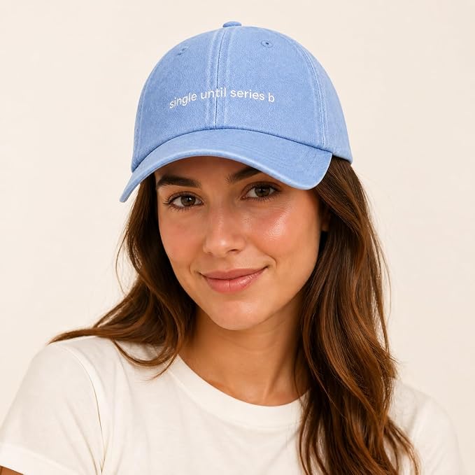 A woman wearing a light blue denim baseball cap with white embroidered text reading 'single until series b' on the front, paired with a white t-shirt against a plain cream background.