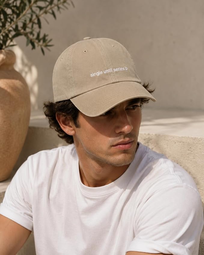 A man wearing a beige baseball cap with white embroidered text reading 'single until series 5' and a white t-shirt, posed in a warm, sunlit outdoor setting.