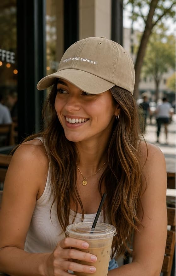 A woman smiling at an outdoor cafe while wearing a beige baseball cap with white embroidered text that reads 'anything but series b'. She is holding an iced coffee.