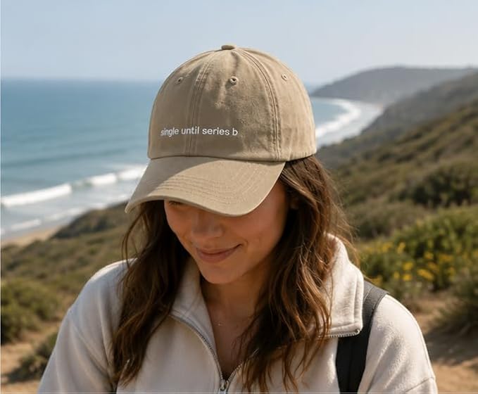 A woman wearing a beige baseball cap with the text 'single until series b' in white, photographed outdoors against a coastal hillside and ocean background.