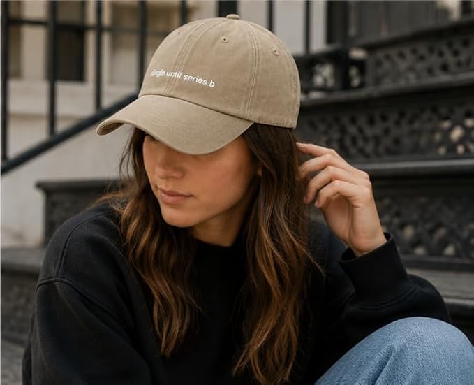 A woman wearing a beige baseball cap with white embroidered text that reads 'single until series b' and a black sweatshirt, sitting outdoors against a black wrought-iron staircase.