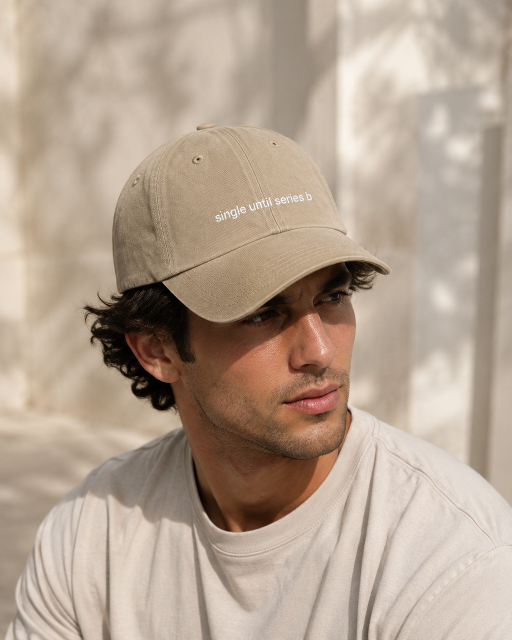 A man wearing a beige baseball cap with white embroidered text reading 'single until series b' and a light beige t-shirt, set against a softly lit, textured beige background.