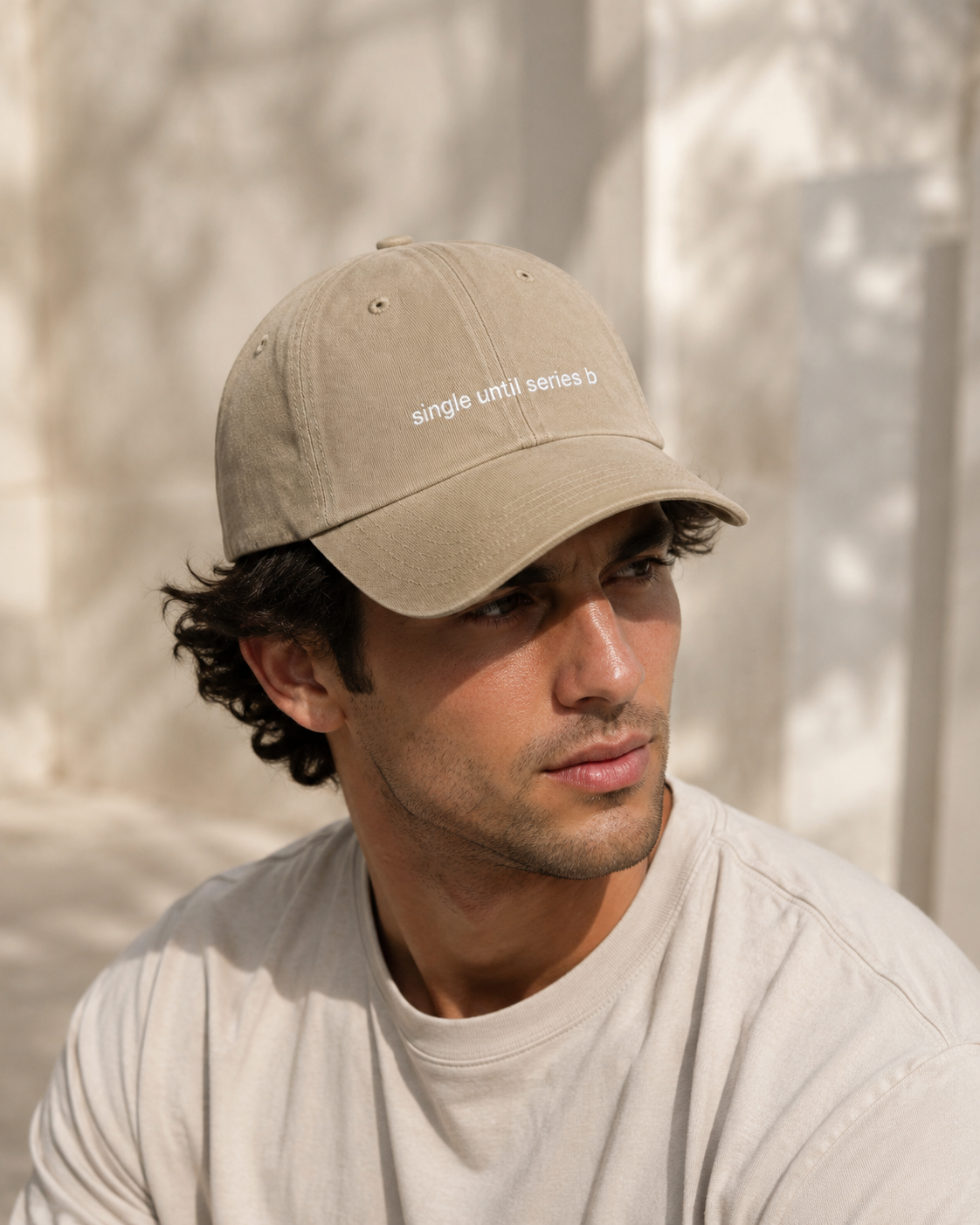 A man wearing a beige baseball cap with white embroidered text reading 'single until series b' and a light beige t-shirt, set against a softly lit, textured beige background.