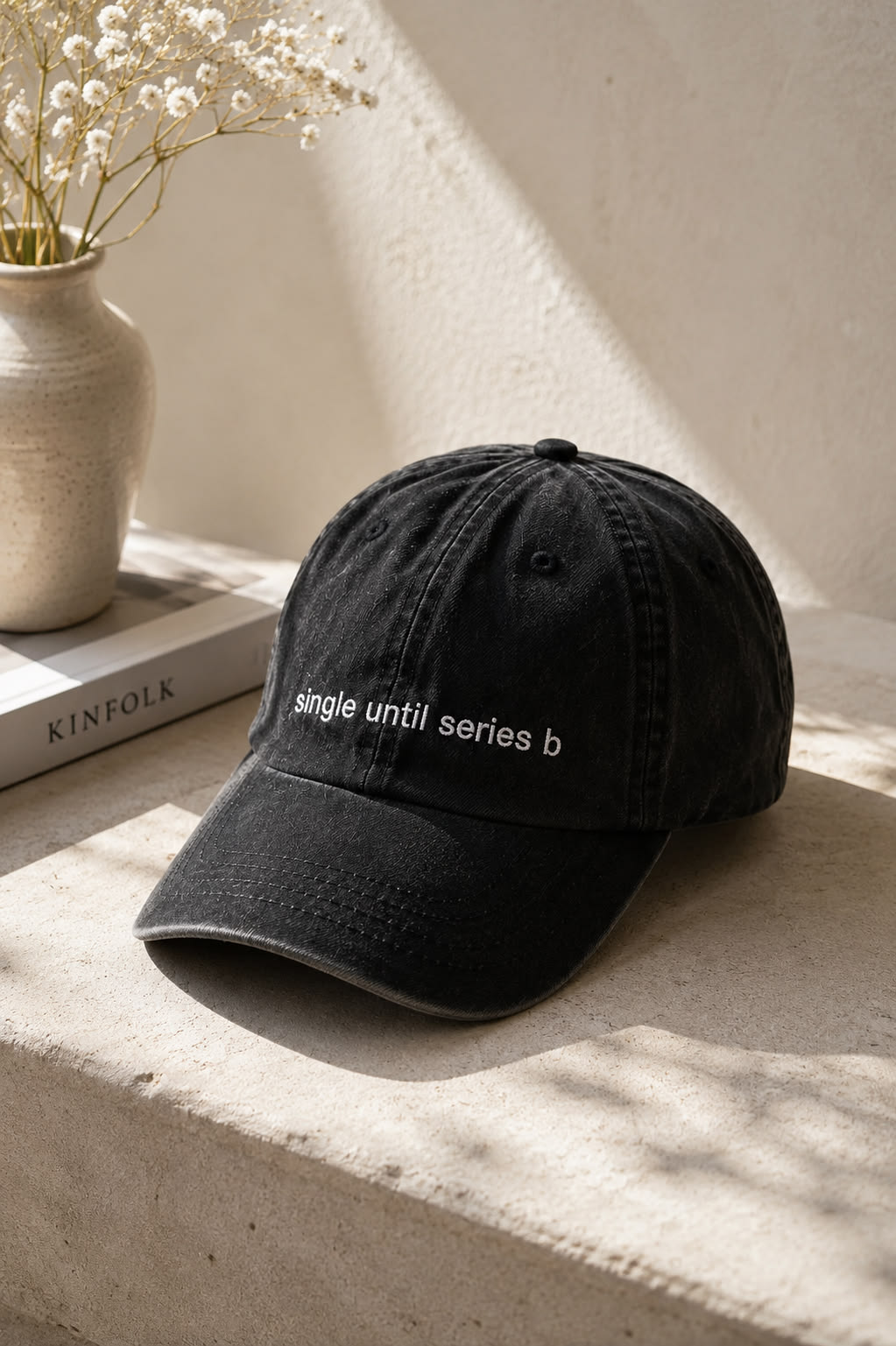 A black baseball cap with white embroidered text reading 'single until series b' resting on a textured stone ledge next to a ceramic vase with dried flowers.