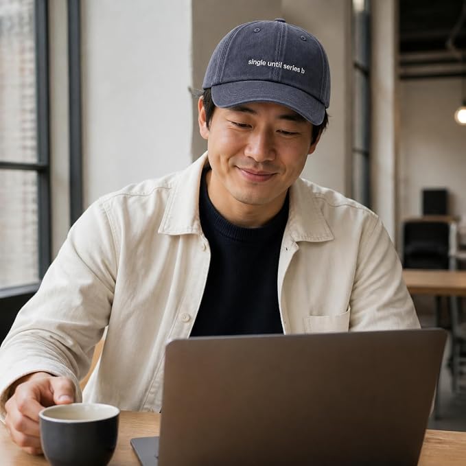 A man wearing a navy blue baseball cap with the embroidered text 'single until series 5' sits at a desk using a laptop and holding a coffee cup in a bright office setting.