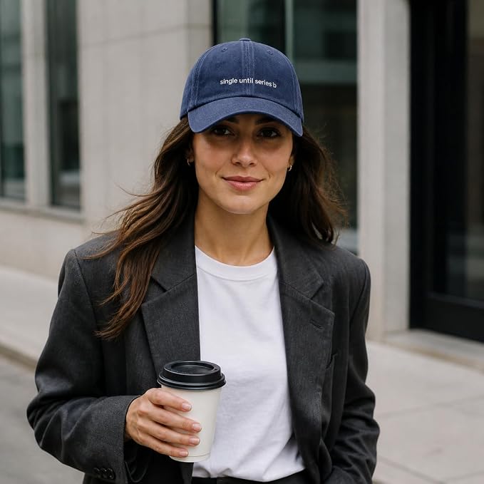 A woman wearing a navy blue baseball cap with white embroidered text that reads 'single until series 3' stands outdoors in front of a modern building, holding a white coffee cup.