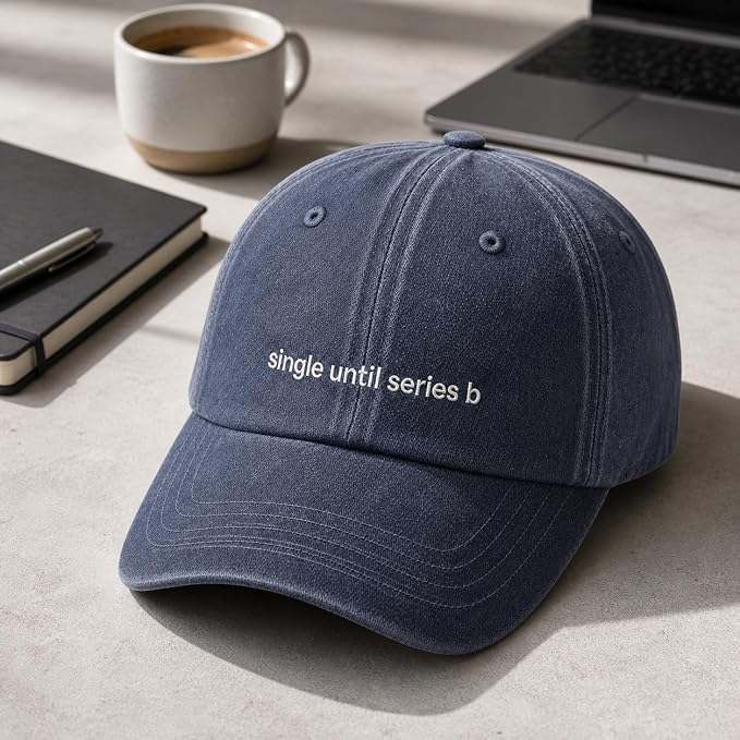 A navy blue washed cotton baseball cap with the phrase 'single until series b' embroidered in white on the front, displayed on a light gray desk with a laptop, notebook, and coffee cup in the background.