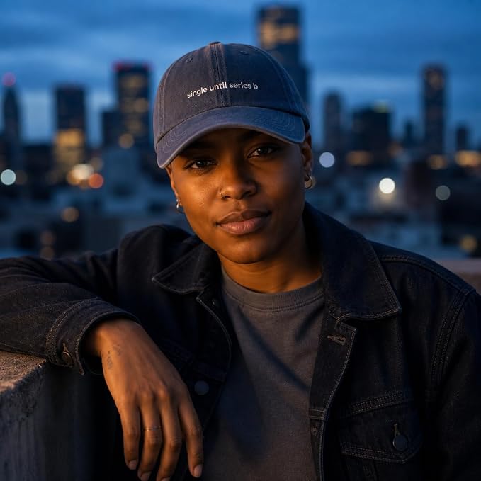 A person wearing a navy blue baseball cap with the white embroidered text 'single until series is' and a dark denim jacket, leaning on a railing with a blurred city skyline at dusk in the background.