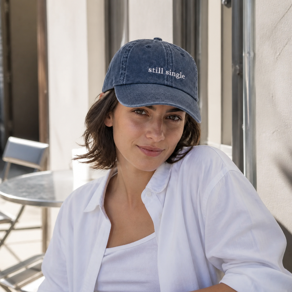 A person wearing a navy baseball cap embroidered with 'still single' in white, shown in profile.