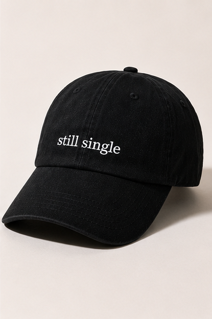 A black cotton baseball cap with the phrase 'still single' embroidered in white lowercase letters on the front, set against a plain light beige background.