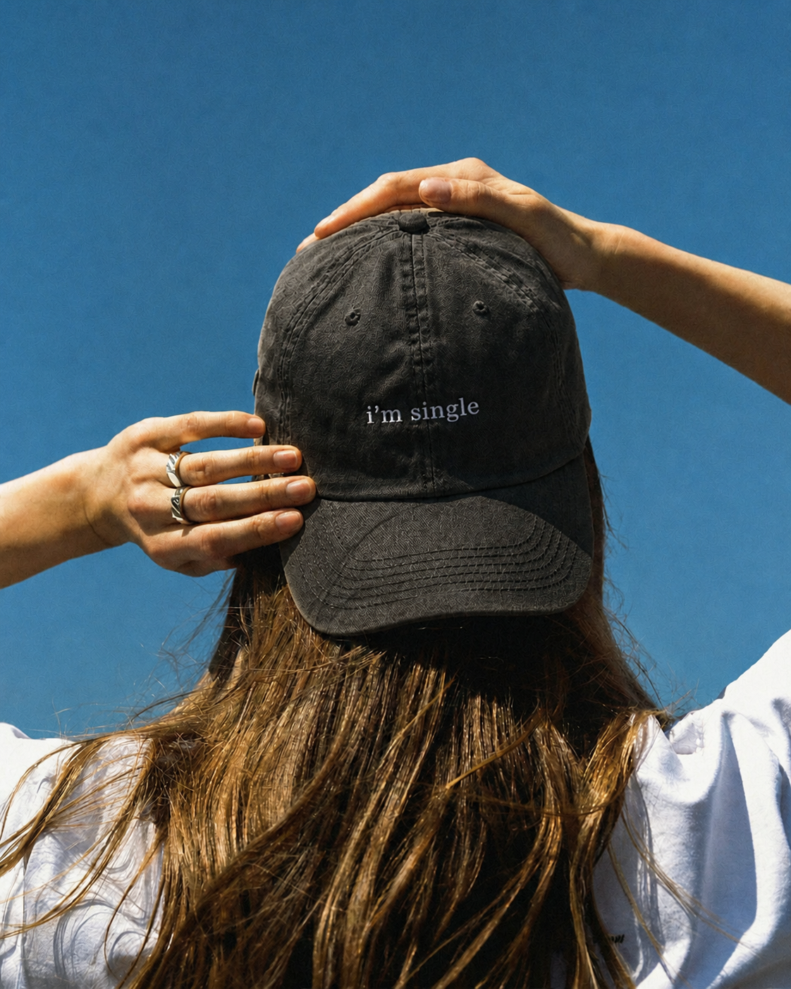 A person viewed from behind wearing a black baseball cap with the text 'i'm single' embroidered in white on the front, set against a clear blue sky.