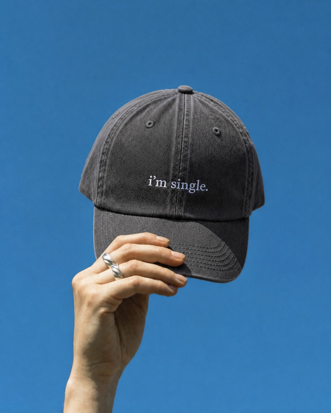 A dark gray washed baseball cap with the phrase 'i'm single.' embroidered in white on the front, held by a hand against a solid blue sky background.